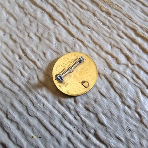 May include: The back of a round, gold-colored pin with a pin back and the letter "D" engraved on the surface.