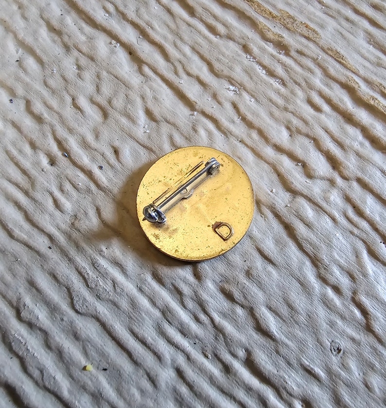 May include: The back of a round, gold-colored pin with a pin back and the letter "D" engraved on the surface.
