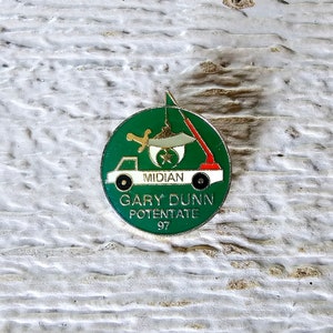 May include: A green and white round pin with a truck carrying a crescent moon and star. The text on the pin reads "Midian Gary Dunn Potentate 97".