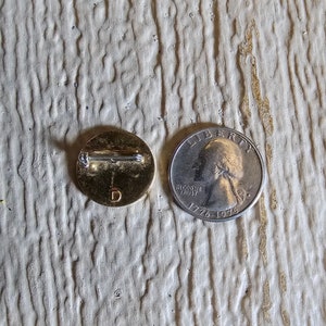 May include: A gold-colored pin with a letter "D" on the front. The pin is next to a US quarter with the year 1976 on it.
