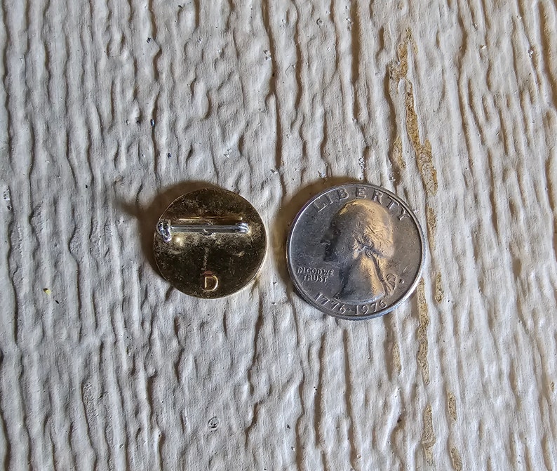 May include: A gold-colored pin with a letter "D" on the front. The pin is next to a US quarter with the year 1976 on it.