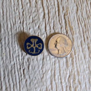 May include: A gold-colored Girl Scout pin with a blue enamel center featuring the iconic trefoil logo. The pin is next to a US quarter for size comparison.