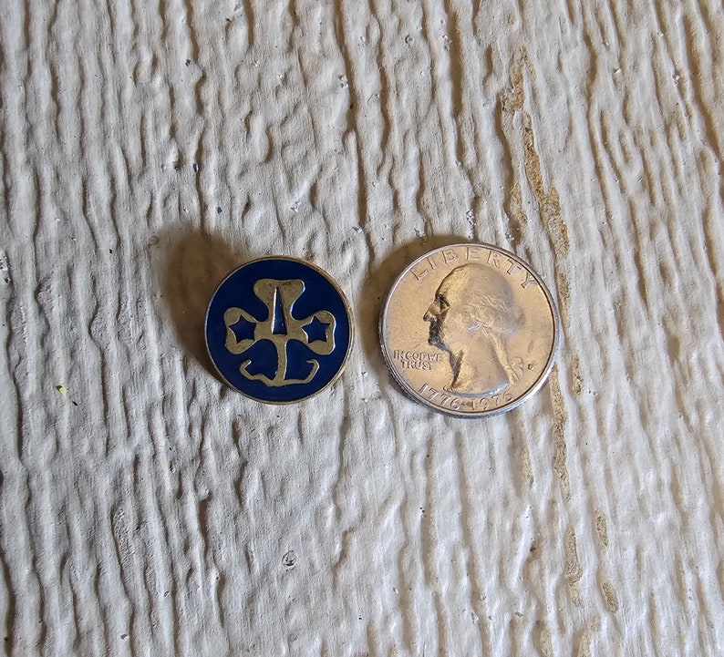 May include: A gold-colored Girl Scout pin with a blue enamel center featuring the iconic trefoil logo. The pin is next to a US quarter for size comparison.