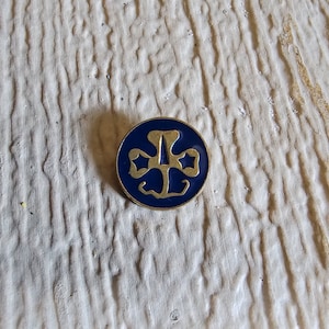 May include: A gold-colored pin with a blue enamel Girl Scout logo featuring a three-leaf clover with a star in each leaf.