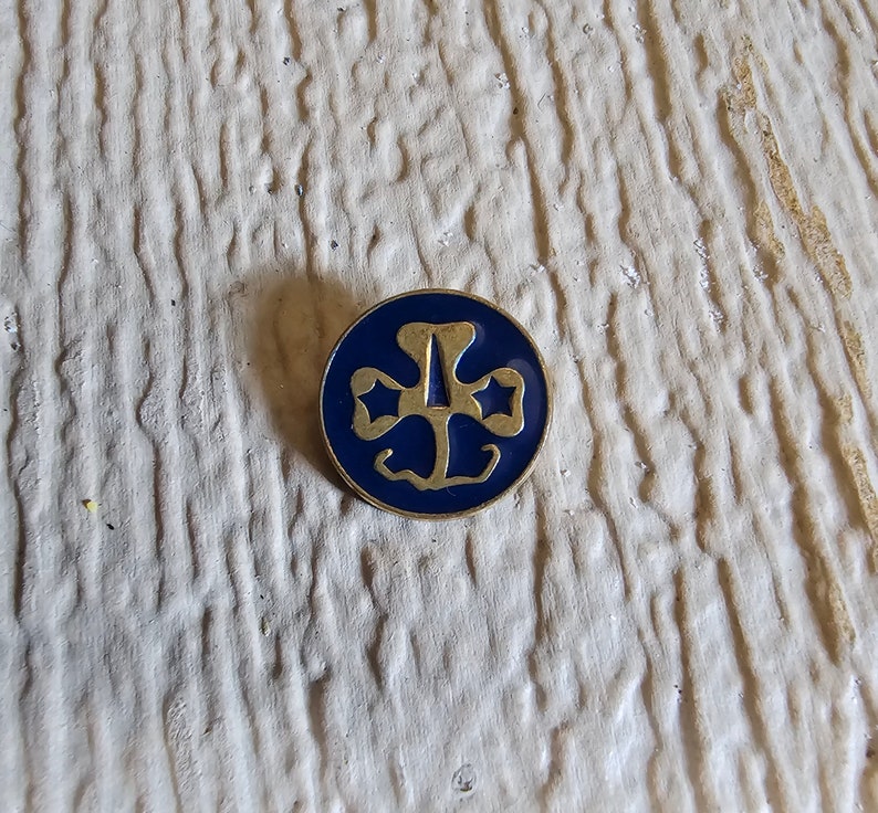 May include: A gold-colored pin with a blue enamel Girl Scout logo featuring a three-leaf clover with a star in each leaf.