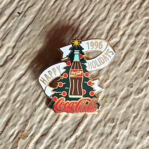 May include: Gold-tone pin featuring a Coca-Cola bottle on a Christmas tree with a banner that reads "Happy Holidays 1996".