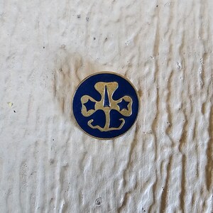 May include: A gold-colored Girl Scout pin with a blue enamel background. The pin features a three-leaf clover with a star in each leaf and a stylized arrow in the center.