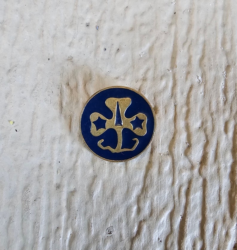 May include: A gold-colored Girl Scout pin with a blue enamel background. The pin features a three-leaf clover with a star in each leaf and a stylized arrow in the center.