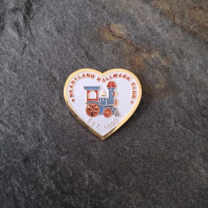 May include: Heart-shaped pin with a gold border and a white center. The pin features a red, blue, and white train design with the text "Heartland Hallmark Club" and "Est. 1990".