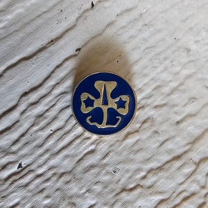 May include: A gold-colored Girl Scout pin with a blue enamel background. The pin features the iconic Girl Scout trefoil logo with three stars.
