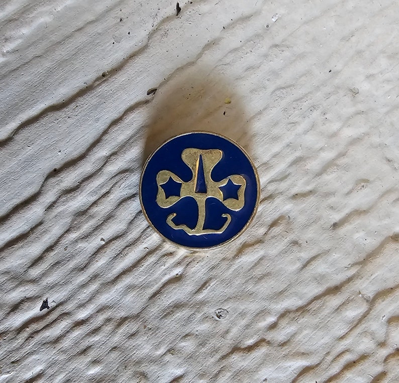 May include: A gold-colored Girl Scout pin with a blue enamel background. The pin features the iconic Girl Scout trefoil logo with three stars.