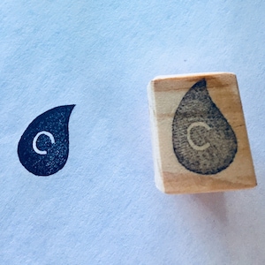 May include: A wooden stamp with a black ink impression of a teardrop shape with the letter "C" inside. The stamp is on a light blue surface.
