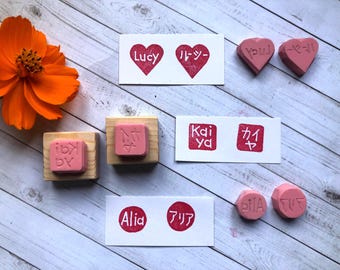 May include: A collection of pink rubber stamps with wooden handles, alongside stamped impressions on white cards. The stamps feature names like "Lucy," "Kaiya," and "Alia" in English and Japanese. A vibrant orange flower adds a pop of color.