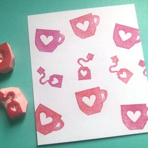 May include: Pink rubber stamps with heart-shaped cutouts next to a white paper with a repeating pattern of pink teacups, tea bags, and hearts. The stamps are used to create the pattern on the paper.