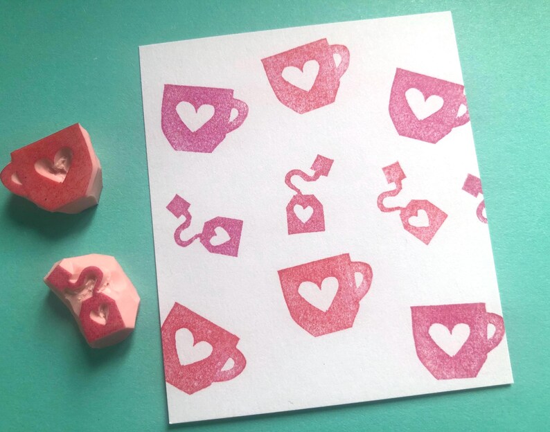 May include: Pink rubber stamps with heart-shaped cutouts next to a white paper with a repeating pattern of pink teacups, tea bags, and hearts. The stamps are used to create the pattern on the paper.