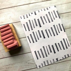 Puede incluir: Un sello de goma rosa con una base de madera junto a un papel blanco con un patrón de líneas verticales negras repetidas. El sello tiene un diseño estriado. El papel está sobre una superficie de madera clara.