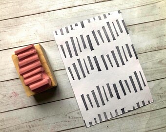 Könnte beinhalten: Ein rosa Gummistempel mit einem geriffelten Muster auf einem Holzsockel, neben einem weißen Papier mit einem sich wiederholenden Muster aus schwarzen vertikalen Linien. Die Artikel befinden sich auf einer hellen Holzmaserung.