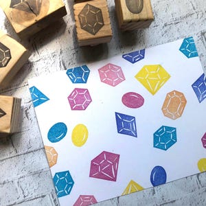 May include: A white sheet of paper featuring a repeating pattern of stamped colourful gemstones in shades of blue, pink, yellow, and orange, accompanied by four wooden rubber stamps with matching geometric gem designs on their faces.