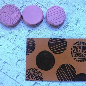 Puede incluir: Tres sellos circulares rosas con diferentes patrones geométricos se muestran encima de una tarjeta marrón. La tarjeta presenta un patrón de círculos negros, algunos con diseños de rayas o entrecruzados, creando un contraste.