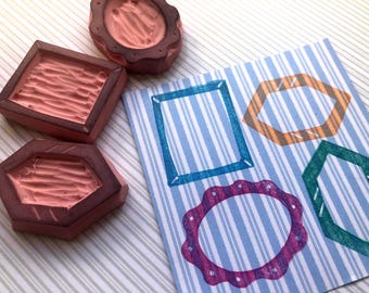May include: Three rubber stamps with carved peach-colored surfaces and dark brown bases, positioned next to a square paper featuring four stamped frame designs in blue, orange, green, and purple on a light blue and white vertical striped background.