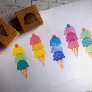 May include: Three wooden rubber stamps with geometric designs are positioned next to a white sheet of paper featuring five stamped illustrations of multi-colored ice cream cones in various pastel and vibrant hues, including pink, yellow, blue, and green.