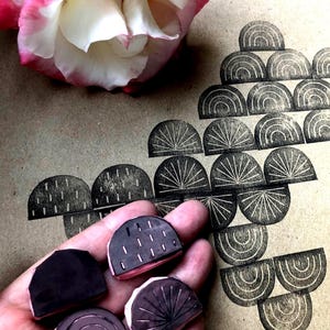 May include: A collection of handmade rubber stamps with geometric designs, including arches and sunburst patterns. The stamps are in various shapes and sizes, with a pink and black color scheme. A pink and white rose is in the background.