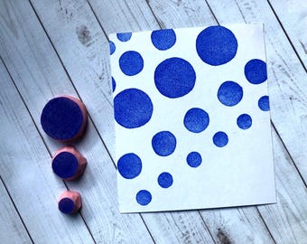 May include: Three circular rubber stamps in varying sizes with pink bases and blue ink on the faces, positioned next to a white rectangular card featuring a scattered pattern of blue circles of different sizes stamped onto the surface.