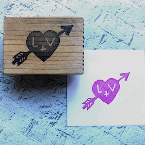 May include: A rectangular wooden rubber stamp featuring a heart pierced by an arrow with the letters L+V engraved on the surface. Next to the stamp is a white square paper showing a purple ink impression of the same heart and arrow design.