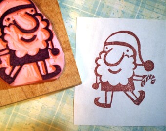 May include: A Santa Claus rubber stamp with a cartoon design, next to a stamped image of Santa. The stamp is pink and black, mounted on a wooden block. The stamped image is red on white paper, Santa is holding a candy cane.