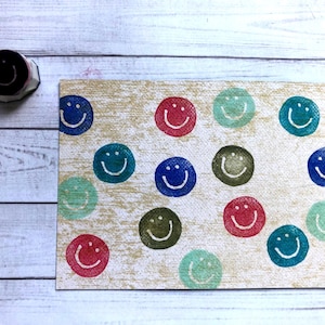 May include: A rubber stamp with a smiling face next to a card printed with multiple colorful smiley faces. The smiley faces are in red, blue, green, and olive green on a beige background.