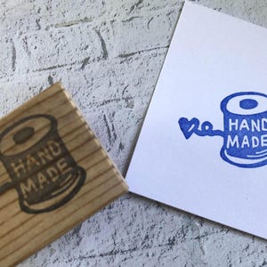 Puede incluir: Un sello de goma con base de madera que muestra un carrete de hilo con el texto HAND MADE y un pequeño corazón, junto a una tarjeta de papel blanco con una impresión en tinta azul del mismo diseño, mostrando el detalle del sello.