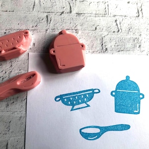 May include: Three pink rubber stamps shaped like a colander, a lidded pot, and a spoon. Next to the stamps is a white sheet of paper featuring blue ink impressions of the same three kitchen utensil designs, showing a colander, a pot, and a spoon.