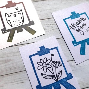 以下が含まれることがあります： 手書きの絵が描かれたカードが3枚あります。最初のカードには、茶色と黒の絵で描かれたフクロウがイーゼルに描かれています。2枚目のカードには、青い絵で「ありがとう」という言葉がイーゼルに描かれています。3枚目のカードには、青い絵で花がイーゼルに描かれています。