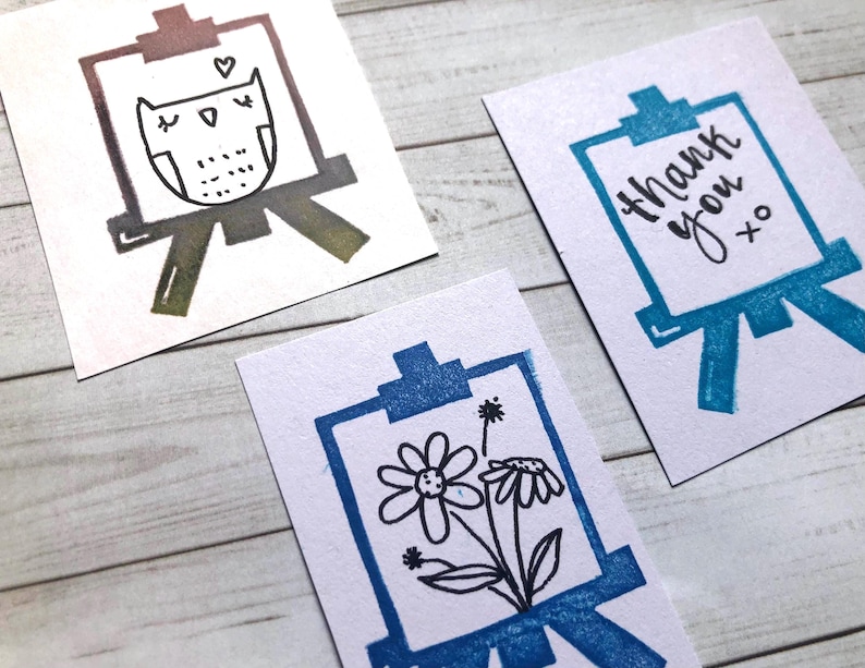 以下が含まれることがあります： 手書きの絵が描かれたカードが3枚あります。最初のカードには、茶色と黒の絵で描かれたフクロウがイーゼルに描かれています。2枚目のカードには、青い絵で「ありがとう」という言葉がイーゼルに描かれています。3枚目のカードには、青い絵で花がイーゼルに描かれています。