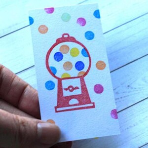 May include: A white card with a red gumball machine illustration and colorful polka dots. The gumball machine has a round glass area filled with yellow, orange, and blue circles. The card is held by a hand.