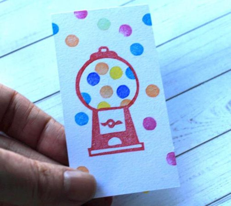 May include: A white card with a red gumball machine illustration and colorful polka dots. The gumball machine has a round glass area filled with yellow, orange, and blue circles. The card is held by a hand.