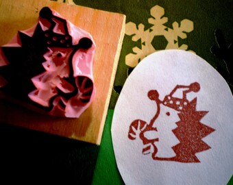 May include: A festive Christmas rubber stamp. The stamp depicts a whimsical character with a Christmas tree body, a candy cane, and a Santa hat. The stamp is mounted on a wooden block, and a stamped image is visible on a white oval. The image is in red.