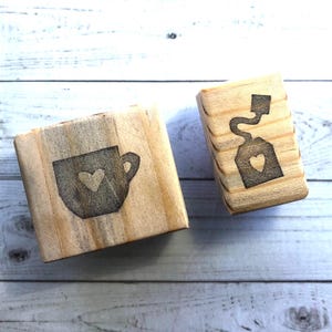 May include: Two wooden blocks with engraved designs. One block features a teacup with a heart, the other a teabag with a heart. The blocks are made of light-colored wood and are likely used for crafting or decoration.