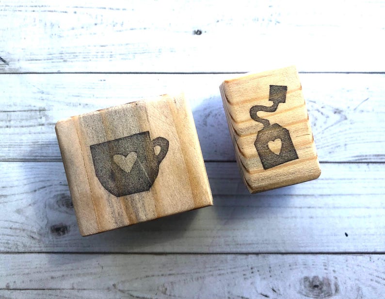 May include: Two wooden blocks with engraved designs. One block features a teacup with a heart, the other a teabag with a heart. The blocks are made of light-colored wood and are likely used for crafting or decoration.