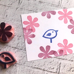 May include: A pink rubber stamp with a dark purple flower design and a second stamp with a bird design. A white card features a blue bird illustration and pink flower border.