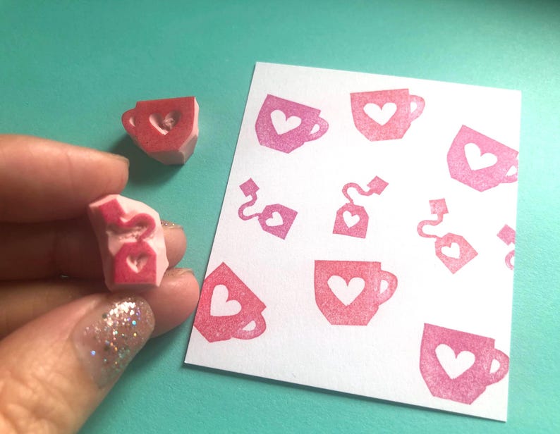 May include: Pink rubber stamps with a teacup and heart design. One stamp is held in a hand, showing the carved teacup shape. A white card displays a pattern of stamped teacups and tea bags with hearts.