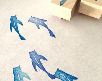 May include: Two rectangular wooden blocks with rubber stamps on the bottom, each featuring a blue fish design. Below the blocks, four blue fish shapes are stamped onto the surface, showing a textured, gradient blue ink pattern for each fish.