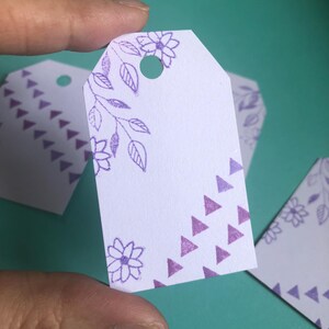 以下が含まれることがあります： 紫色のギフトタグ。花柄と紫色の三角形の模様が施されています。タグの上部に紐やリボンを通すための穴が開いています。