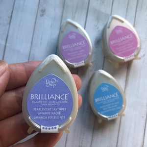 May include: Four pearlescent pigment ink dew drops in lavender, pink, and blue. The teardrop-shaped containers have the word "BRILLIANCE" printed on them. The ink is labeled as "Pearlescent Lavender" and "Lavande Nacrée".