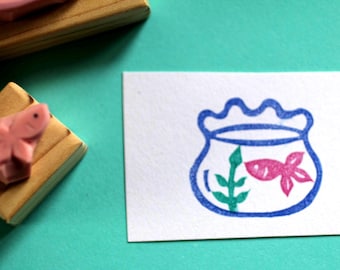 May include: A blue and white rubber stamp with a design of a fishbowl with a single goldfish inside. The fishbowl has a green plant inside. The stamp is on a white card.