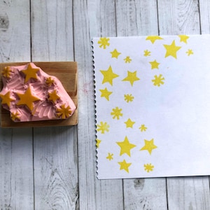 May include: A pink rubber stamp with a star design and a wooden base. The stamp has several yellow stars of varying sizes. A white card with yellow star imprints is next to the stamp. The background is a light wood surface.