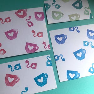 May include: Four white cards with a repeating pattern of tea cups and tea bags, each with a heart in the center. The cups and tea bags are in shades of pink, blue, and green. The cards are arranged on a teal surface.