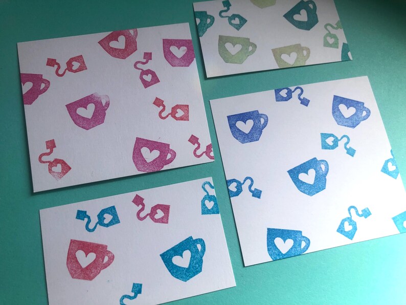 May include: Four white cards with a repeating pattern of tea cups and tea bags, each with a heart in the center. The cups and tea bags are in shades of pink, blue, and green. The cards are arranged on a teal surface.