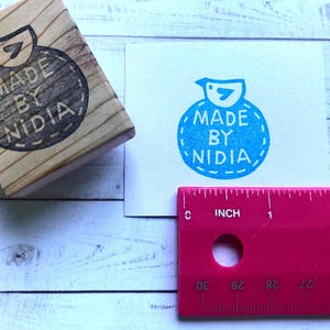 以下が含まれることがあります： 青い鳥と「MADE BY NIDIA」という文字が縫い付けられた縁取りのある円の中に書かれた木製のスタンプ。スタンプは白い木製の表面の上に置かれています。