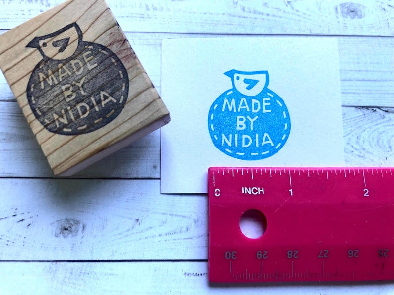 以下が含まれることがあります： 青い鳥と「MADE BY NIDIA」という文字が縫い付けられた縁取りのある円の中に書かれた木製のスタンプ。スタンプは白い木製の表面の上に置かれています。